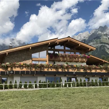 A modern, well-designed house with a balcony full of flowers. In the background, majestic mountains and a blue sky with some clouds can be seen.