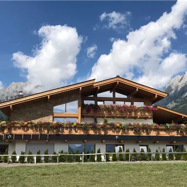 A modern alpine house with a wooden facade and colorful flowers. Impressive mountains and a blue sky are visible in the background.