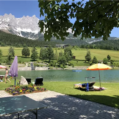 Ein schöner See umgeben von grünen Wiesen und Bergen. Schwimmer genießen das Wetter unter Sonnenschirmen.