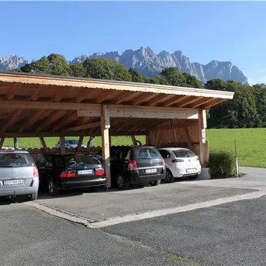 A parking lot with several cars under a wooden carport. In the background, green meadows and mountains can be seen.