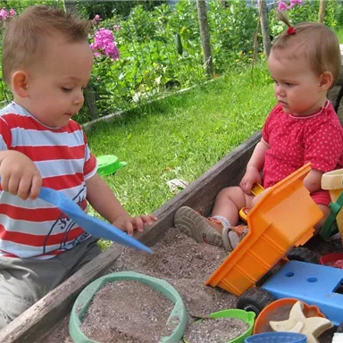 Zwei kleine Kinder spielen im Sandkasten. Der Junge gräbt mit einer Schaufel, während das Mädchen daneben sitzt und Spielzeug um sich hat.