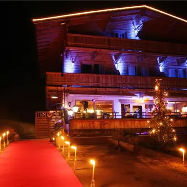 A cozy mountain chalet at night, illuminated with colorful lights. A red carpet leads to the front door, surrounded by torches and festive decorations.