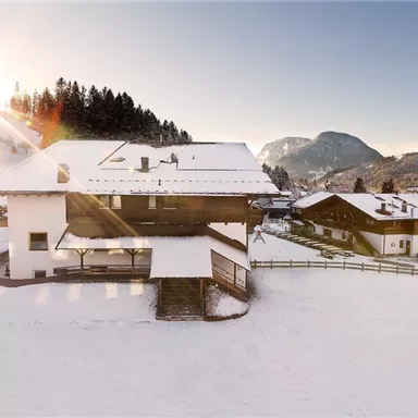 A beautiful winter landscape with snow-covered houses and a clear blue sky. The sun shines over the mountains, giving the scene a picturesque atmosphere.