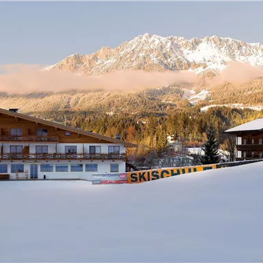 A snowy mountain landscape with two chalet houses. Snow-covered mountains and a clear sky stretch out in the background.