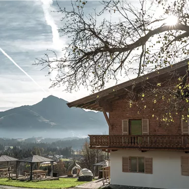 Ein traditionelles Holzhaus liegt malerisch in einer ländlichen Umgebung. Im Hintergrund erhebt sich ein hoher Berg unter einem klaren Himmel.
