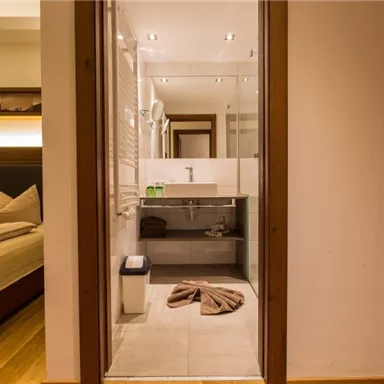 A modern bathroom with a glass shower and light tiles. In the background, a cozy bedroom with a large bed can be seen.