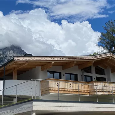 A modern house with a covered balcony and wooden details. In the background, mountains and a cloudy sky can be seen.