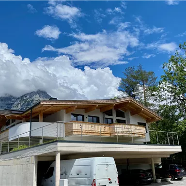 A modern house with a balcony and a carport stands in a green environment. In the background, mountains and a cloudy but sunny sky can be seen.