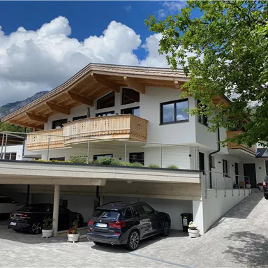 Ein modernes Haus mit Holzbalustrade und einer überdachten Garage. Im Hintergrund sind blühende Bäume und ein blauer Himmel zu sehen.