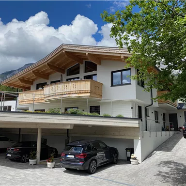 A modern house with a large porch and wooden details. In the foreground, several cars can be seen on a paved parking lot.