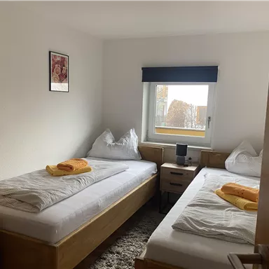 Ein einfaches Schlafzimmer mit zwei Einzelbetten und weißen Bettdecken. Auf den Betten liegen orangefarbene Handtücher, und ein Fenster lässt Tageslicht herein.