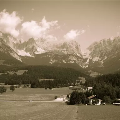 Eine beeindruckende Berglandschaft mit hohen Gipfeln und einem klaren Himmel. Im Vordergrund erstreckt sich eine grüne Wiese und kleine Gebäude.