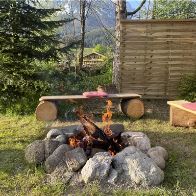 Ein gemütliches Lagerfeuer, umgeben von Steinen und Holzbänken. Im Hintergrund  ist das Haus Gaberhel sichtbar.