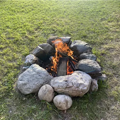 Ein Lagerfeuer in einem Steinkreis auf Grünfläche. Das Feuer brennt hell und erzeugt eine warme Atmosphäre.