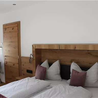 A cozy bedroom with a wooden headboard and two large pillows. The room is bright and modernly furnished, with a small table and chairs in the corner.