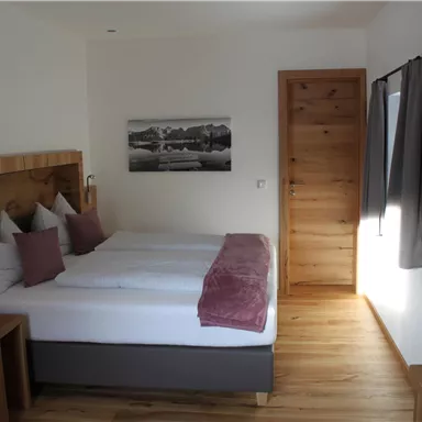 A modern bedroom with a double bed and wooden details. The walls are light, and there is a large window with curtains.