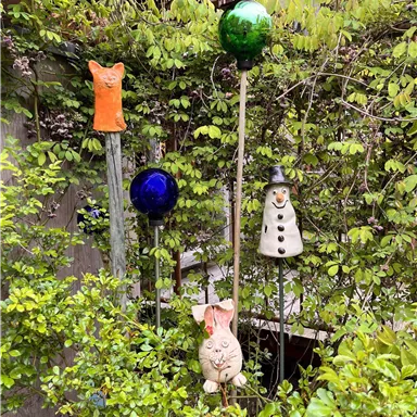 Fünf bunte Gartendekorationen stehen zwischen grünen Pflanzen. Darunter sind Tier- und Figuren aus Keramik, die eine fröhliche Atmosphäre schaffen.