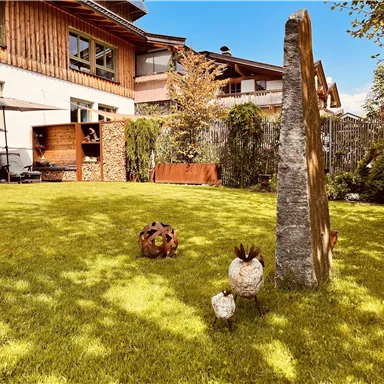 Ein gepflegter Garten mit grünem Rasen und einer Steinskulptur. Im Hintergrund ist ein Haus und einige dekorative Figuren zu sehen.