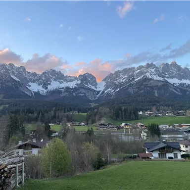 A picturesque mountain panorama with snow-covered peaks and gentle meadows. In the foreground, small houses and a dense forest can be seen.