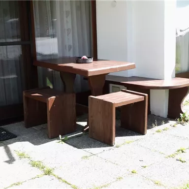 Ein kleiner Außenbereich mit einem Holztablett und zwei Hockern. Die Möbel stehen neben einem Gebäude und sind umgeben von einem gepflegten Rasen.