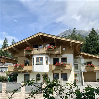 Ein schönes Chalet mit Balkonen, geschmückt mit blühenden Pflanzen. Im Hintergrund sind Berge und ein klarer Himmel zu sehen.