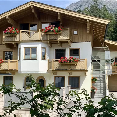 Ein charmantes, mehrstöckiges Holzhaus mit Balkon und blühenden Pflanzen. Im Hintergrund sind Bäume und Berge sichtbar.