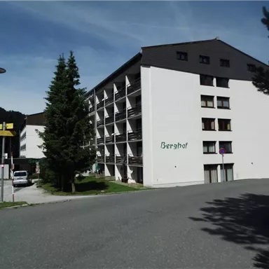 Ein modernes Gebäude namens "Berghof" in einer bergigen Umgebung. Die Eingangstür ist gut sichtbar, und Bäume umgeben die Anlage.