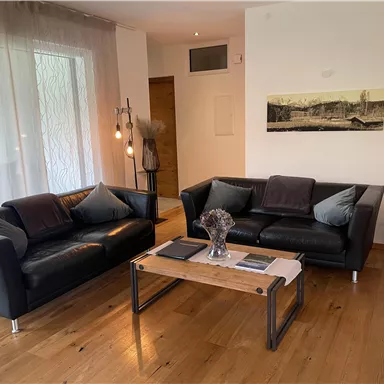 A modern living room with two black sofas and a wooden table. The walls are light, and there are large windows that let in natural light.