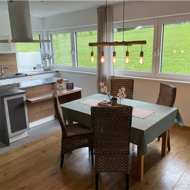 A modern kitchen with a wooden dining area. The table is set and surrounded by chairs with a view of a green meadow.