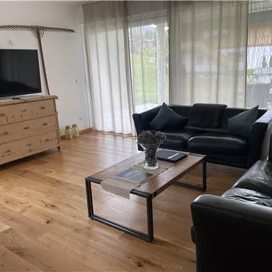 A cozy living room with a television, a wooden cabinet, and two sofas. Large windows let in plenty of light and give the room an inviting atmosphere.
