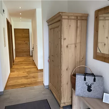 A bright hallway with wooden floors and walls. On the left side, there is a wooden cabinet and a decorative mirror is mounted on the wall.