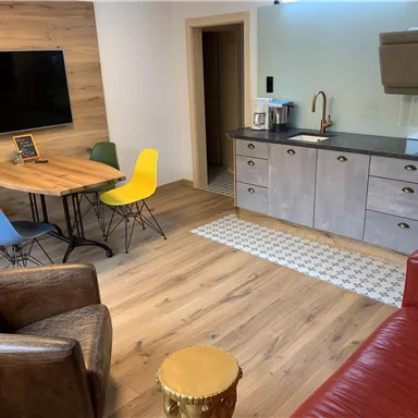 Ein modernes Zimmer mit einem Essbereich und einer Küchenzeile. Bunte Stühle um den Tisch und ein gemütliches Sofa schaffen eine einladende Atmosphäre.