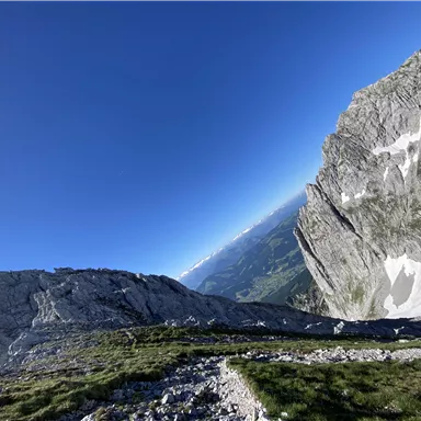 Eine atemberaubende Berglandschaft mit steilen Felsen und einem klaren blauen Himmel. Im Hintergrund sind grüne Täler und weitere Berge sichtbar.
