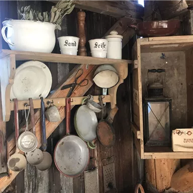 A rustic kitchen with old cooking utensils and dishes. Various pots and an oil lamp hang on the wall.