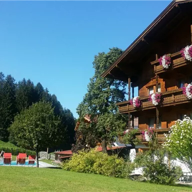 A cozy wooden house with flower boxes on the balconies. In the foreground, a green meadow stretches out with loungers and a view of the wooded mountains.