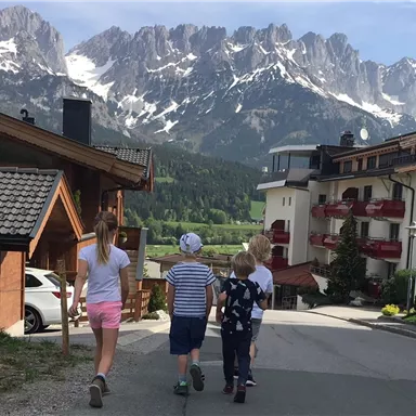 A group of children walks along a street, surrounded by mountains. In the background, houses and a beautiful landscape can be seen.
