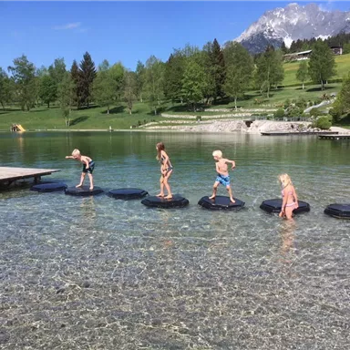 Children are playing on large floating platforms in a clear lake. In the background, trees and mountains can be seen.