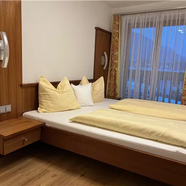 A cozy bedroom with a double bed, wooden paneling, and large windows. The pillows and blankets are in soft colors.
