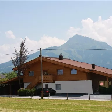 A cozy wooden house with a beautiful mountain in the background. The surroundings are green and the clouds are visible in the sky.