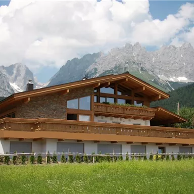 A modern house with wood siding amidst a green meadow. In the background, majestic mountains and a clear sky are visible.