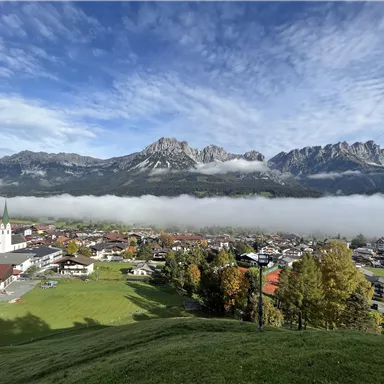 A picturesque landscape with high mountains in the background and a city in the foreground. Mist hovers over the city and the clouds are partially visible.