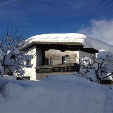 A cozy wooden house, surrounded by towering snow. The sky is clear and blue.