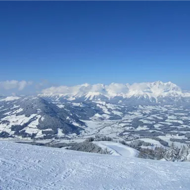 A snow-covered mountain landscape under a clear blue sky. In the distance, majestic mountains and gentle hills can be seen.