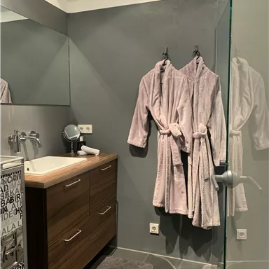 Ein modernes Badezimmer mit einer Holzkommode und zwei flauschigen Morgenmänteln. Der Raum hat große Fliesen und eine Glasduschwand.