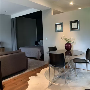 Ein modernes Wohnzimmer mit einem Esstisch aus Glas und schwarzen Stühlen. Im Hintergrund befindet sich ein Schlafzimmer und ein schön dekorierter Teppich.