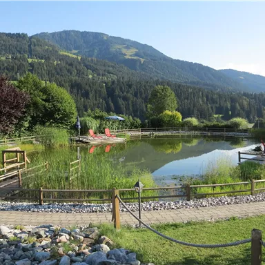 Ein ruhiger Teich umgeben von grünen Wiesen und Bergen. Holzgänge führen zu gemütlichen Sitzbereichen.