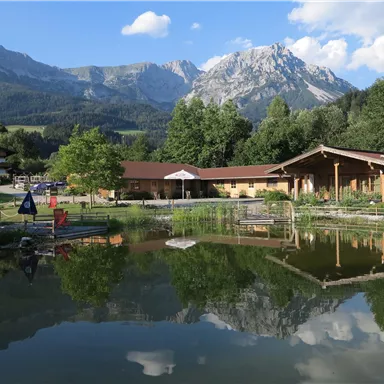 Ein malerisches Bergpanorama mit einem ruhigen Teich im Vordergrund. Die Hütte und die Bäume spiegeln sich im Wasser wider.
