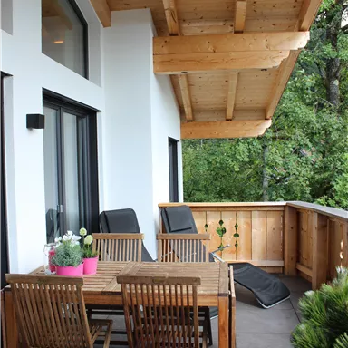 A cozy terrace with a wooden table and wooden chairs. In the background, green trees can be seen.