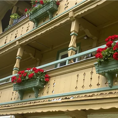 Ein traditionelles Holzgebäude mit einem Balkon, der mit roten Geranien geschmückt ist. Die Architektur zeigt eine detailreiche Fassadengestaltung und bietet einen einladenden Eindruck.
