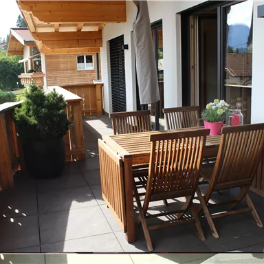 A cozy terrace with a wooden table and chairs. In the background, beautiful mountain landscapes and adjacent houses can be seen.